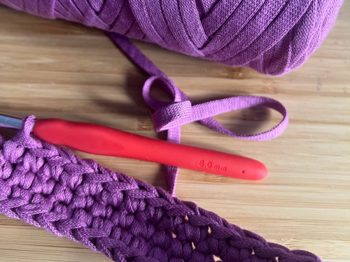 Dettaglio di uncinetto artigianale realizzato con cordino di cotone viola, filato naturale lavorato a mano su piano in legno